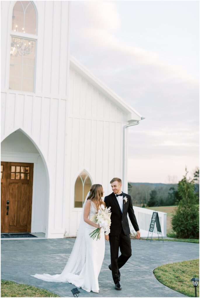 Husband and Wife, Bride and Groom walk hand in hand holding her wedding bouquet in front of their wedding venue