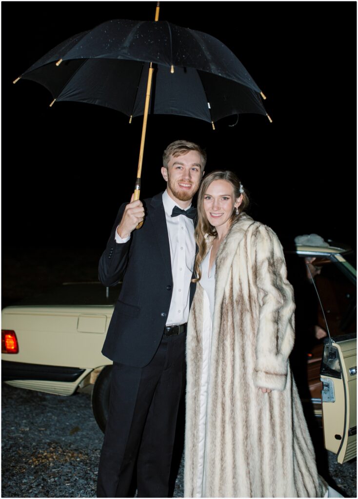 The final exit where the bride and groom are standing in front of old car holding an umbrella after their wedding