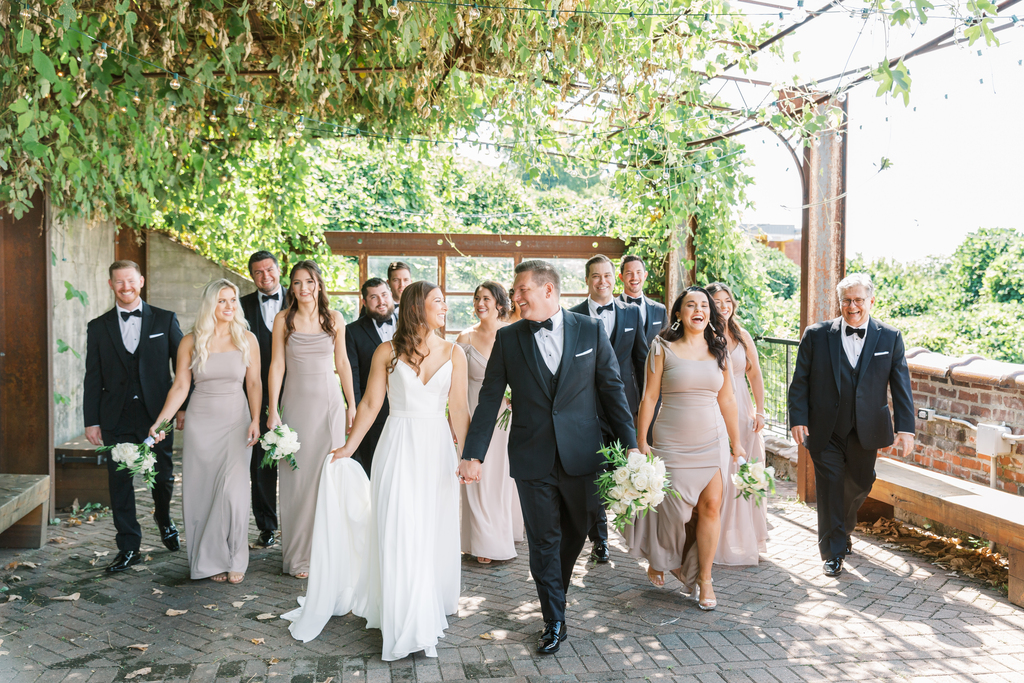 Downtown wedding with bridesmaids and groomsmen walking and laughing in photo, the bride and groom are holding hands and are in front of the large group walking.